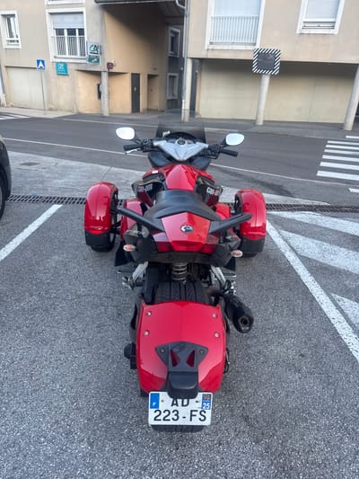 In the city of Lavans-lès-Saint-Claude, France, a red three-wheeled Can-Am Spyder motorcycle is parked on an asphalt surface marked with white lines. The motorcycle is positioned centrally in the frame, viewed from the rear. It features a black seat, chrome accents, and a visible license plate that reads "AD 223-FS" with a French emblem and the number 25. To the left of the motorcycle, a portion of a black vehicle is visible. In the background, a building with beige walls and windows is present. A pedestrian crossing is visible to the right of the motorcycle.