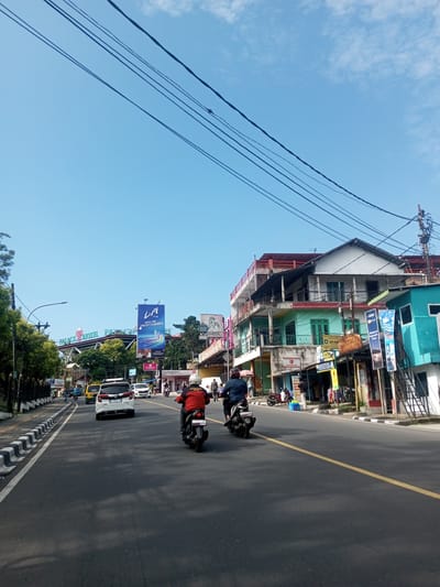 The image depicts a street scene in Khe Luang, Laos. The photo is taken from a ground-level perspective, looking down a road with traffic. Two motorbikes are traveling in the same direction as the camera, along with a white car and a yellow vehicle. The road has a single yellow line marking the center. Buildings line the right side of the street, with a multi-story building in the mid-ground featuring turquoise and white paint. On the left side of the street, there is a sidewalk and trees. In the background, signs are visible, including one for the "Palace Hotel." Overhead, there are power lines stretching across the sky. The sky is blue with some cloud cover.