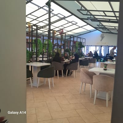 The image shows the interior of a cafe named "Sky View," located in Al-Mawasi. The cafe has a modern design with a grid-like structure forming the ceiling, interspersed with hanging plants. Several people are seated at tables, some of them engaged in conversation. The furniture consists of light-colored tables and chairs. The floor is tiled in a light brown pattern. A "Galaxy A14" watermark is visible at the bottom right corner of the image.