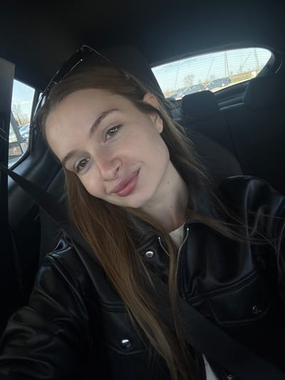 A woman is seated in a car, wearing a black leather jacket and a seatbelt. She has long brown hair, light skin, and is looking towards the camera. Sunglasses are perched on her head. The scene suggests the car is parked; outside the car window, buildings and other vehicles can be seen. The reflection of the environment suggests a cityscape, possibly of Ub Municipality, Serbia.