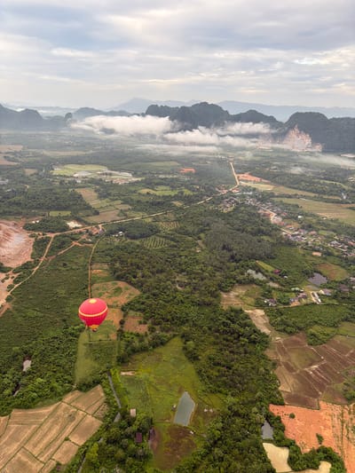 An aerial shot captures a red hot air balloon with yellow accents floating over a lush green landscape in Hong Ngern Village, Laos. The village is situated amidst rolling hills and dense forestation, with rice paddies and small bodies of water dotting the terrain. In the background, a mountain range rises, partially shrouded in mist, with a cloudy sky overhead. The scene depicts a serene and natural environment, likely taken during a morning balloon ride.