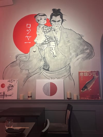 A wall mural depicts a sumo wrestler embracing a child against a red sun. Japanese text complements the artwork. Above, a retro advertisement featuring a fish is displayed. Adjacent to the mural, a series of framed prints includes a minimalist sun over water design and a bamboo illustration, all punctuated by small, tied candles. A dark chair and table with tableware occupy the right side, adding a contemporary element to the setting.