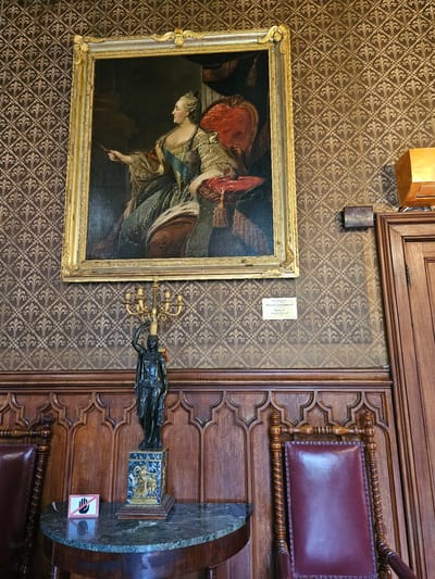 A photograph shows a framed painting hanging on a patterned wall, alongside a decorative wooden panel and a statue on a marble-topped table, likely taken in a historical or cultural institution.
The painting, set in a gold frame, depicts a woman in a historical dress. To the right of the painting, a bronze statue of a woman holding a candelabra is placed on a pedestal on a table with a marble top. A "Do Not Touch" sign is visible on the table. The wooden panel behind the table features ornate carvings and a leather-upholstered seat.
The wallpaper has a repeating decorative pattern in shades of brown and gold. A small electrical box is visible above the painting. The location is possibly in Алупкинский городской совет, Ukraine, based on the file name metadata.