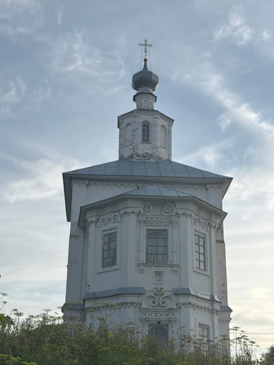 The image shows a close-up, low-angle view of a white, multi-tiered Orthodox church tower against a cloudy sky. The tower has octagonal sections with arched windows and ornate architectural details. A dark green dome with a cross tops the structure. In the foreground, dense green vegetation, including tall plants with umbrella-shaped flowers, obscures the lower part of the church. The scene is set in the city of Cherdyn, Russia.