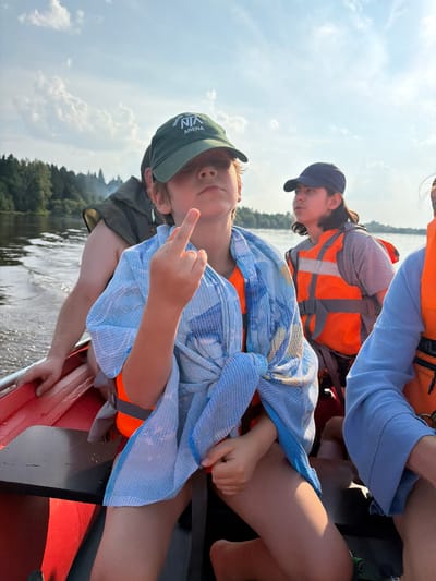 The image shows a group of people in a canoe on a body of water, likely a lake or river. The perspective is from within the canoe, looking up and to the side. Two individuals are prominently featured. One is a young person wearing a green baseball cap and a blue and white patterned towel draped over their shoulders. This person is looking up and making a gesture with their fingers. They are wearing an orange life vest underneath the towel.

Another person is visible behind and slightly above the first, wearing a dark baseball cap and an orange life vest. Their head is tilted down, and they appear to be looking at something. Further in the background, other people in life vests are present, one wearing a blue long-sleeved shirt.

The setting appears to be a natural one, with trees and foliage visible along the distant shoreline. The sky is bright and cloudy, suggesting it is daytime. The water is rippling, indicating movement.

The visible text on the green baseball cap reads "NORTH VIN ARENA". The overall mood seems casual and possibly adventurous, given the context of being in a canoe and wearing life vests. The image is oriented vertically.