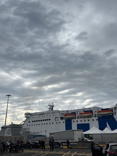 A ferry, identified as "GNV Auriga," docks at Civitavecchia, Italy, under a cloudy sky. Numerous motorcycles park along the dock adjacent to the ferry, with people standing around them. A car sits near the ferry's loading area. The ferry itself is painted white with blue accents.