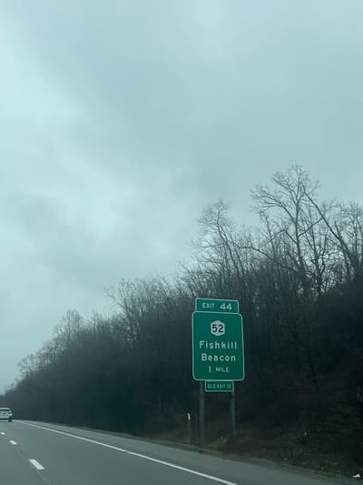 The image shows a highway scene on a cloudy day near the Town of Fishkill, United States of America. A green highway sign indicates "Exit 44" and "52 Fishkill Beacon 1 Mile." A smaller sign below reads "Old Exit 12." Trees line the side of the road, suggesting a somewhat rural or suburban environment. The highway itself is paved with a white lane marker visible. A vehicle is partially visible at the bottom of the picture.