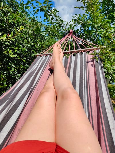 The image shows a person lying in a hammock outdoors. The hammock has a striped pattern of pink, grey, and white. The person's legs and feet are visible, and they are wearing red shorts. The hammock is suspended between two trees, with lush green foliage surrounding it. The sky above is blue with some white clouds, indicating a pleasant day. The sun appears to be shining, casting shadows on the hammock and the surrounding leaves. The overall scene suggests a relaxed and leisurely activity, likely taking place during the daytime in a garden or park. There are no visible signs or text in the image.