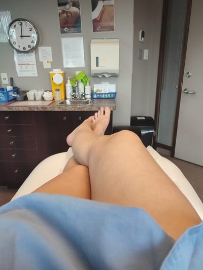 The image shows a first-person perspective from the waist down, with legs crossed and positioned on a white chair or examination table. A blue cloth or sheet is draped over the lower body. In the background, a counter with medical supplies is visible. Items include a yellow sharps container, bottles of soap or sanitizer, a sink with a faucet, and various containers of swabs or cotton balls. A large circular clock hangs on the wall to the left, showing approximately 2:48. Medical posters are also displayed on the wall. To the right, a closed door with a window is seen, and a trash can is nearby. The environment appears to be a medical or clinic setting, potentially a dermatology or aesthetic clinic, given the presence of medical supplies and posters related to skin treatments. The image was taken in Boucherville, Canada.