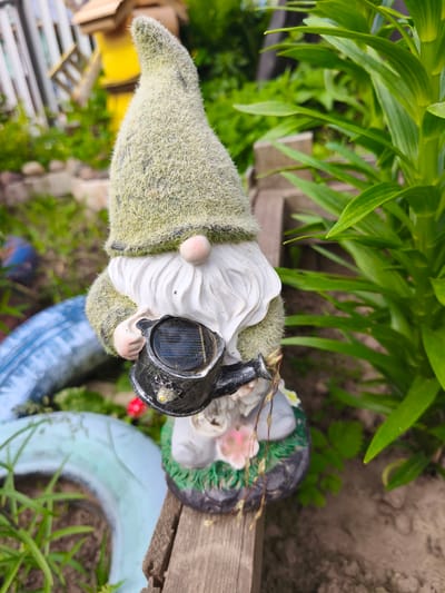 In an outdoor setting, a garden gnome figurine stands prominently on a wooden surface. The gnome, wearing a fuzzy green pointed hat and a white beard, is holding a small black watering can that features a solar panel and a light. The gnome is positioned in front of lush green foliage. In the background, a white wooden fence and a yellow structure are visible. The ground is covered with dirt and some discarded tires, one of which is painted blue and light green. The scene appears to be a garden or yard. The specific location within the city of Puchezh, Russia, is not discernible from the image.