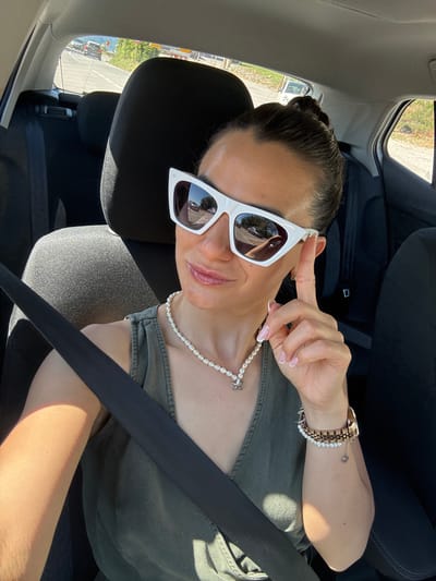 A woman with her hair pulled back in a bun and wearing white cat-eye sunglasses, a pearl necklace, and a green sleeveless top is taking a selfie in the passenger seat of a car. She is wearing a seatbelt. Her left arm is visible, resting on her lap. On her right wrist, she wears a watch and a pearl bracelet with a small charm. The background shows the interior of a car, with a glimpse of a road and trees through the rear window. The location is Blizikuće, Montenegro.