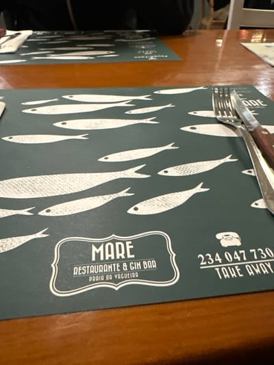 A restaurant placement mat lies on a wooden table. It is decorated with stylized white fish on a dark grey background. It also features the restaurant's name, "Mare Restaurante & Gin Bar," alongside the phone number, "234 047 730," and the phrase "Take Away." Utensils, a fork and a knife, are placed on top of the mat. Another identical placement mat is partially visible to the left.