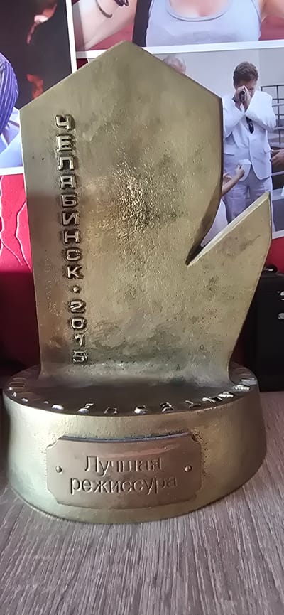 A bronze trophy, likely an award, stands on a wooden surface, possibly a shelf or table. The trophy is predominantly gold-colored with some patina or tarnishing. It features embossed lettering in Russian. The inscription on the side reads vertically: "СЕЗОНА • 2015" which translates to "SEASON • 2015". On the base of the trophy, there is a rectangular plaque with another Russian inscription: "ЛУЧШАЯ РЕЖИССУРА" which translates to "BEST DIRECTION". The trophy has a distinctive shape with a pointed element. In the background, out of focus, are parts of photographs or posters depicting people in various attire, including formal wear and casual clothing. One of the images shows a man in a white suit. The overall environment appears to be indoors, with the wooden surface suggesting a domestic or exhibition setting. The location is identified as Gostagayevskaya, Russia. There are no people directly interacting with the trophy in the frame.