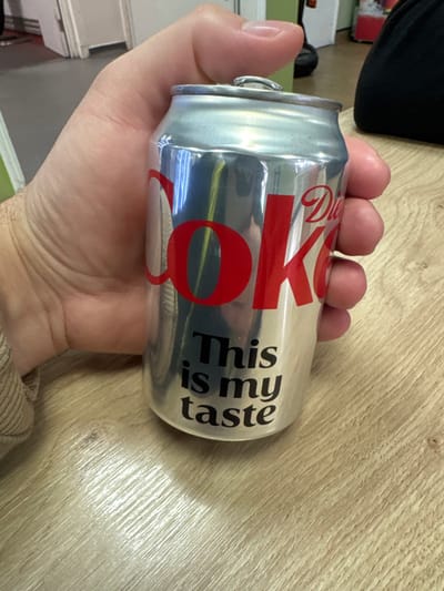 A person is holding a can of Diet Coke in their right hand. The can is silver with red and black lettering. The background is a light brown surface, possibly a wooden table. Other items are visible in the background, out of focus. The location, Basingstoke and Deane, United Kingdom is not discernible from the visual information provided.
