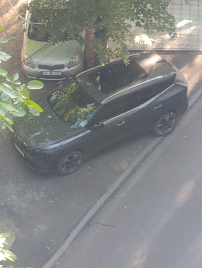 The image shows an elevated view of two cars parked on a street in Soroca, Moldova. The car in the foreground is a dark-colored SUV, possibly black or dark grey, with a sunroof and distinctive alloy wheels. It is parked alongside a paved road with a curb. The second car, a lighter-colored hatchback, is parked behind the SUV and is partially obscured by trees and branches. This vehicle appears to be a Toyota Yaris, identifiable by its headlights and front grille. A license plate reading "SR BN 407" is visible on the front of the hatchback. The scene is set outdoors, with sunlight filtering through the leaves of nearby trees, casting dappled shadows on the ground and cars. A decorative wall with a textured pattern is visible in the background on the right side of the image. The overall environment suggests a residential or street parking area.