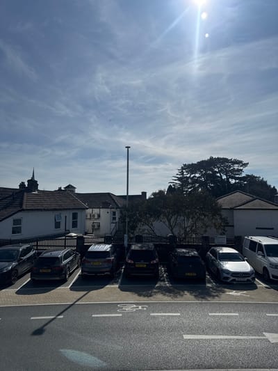 The image depicts a sunlit urban scene in Salisbury, United Kingdom, featuring a parking area, buildings, and sky.
The upper portion of the frame shows a bright blue sky with sparse, wispy cirrus clouds. The sun is visible in the upper right quadrant, emitting bright rays and causing lens flare.
The midground is dominated by a parking area, where seven vehicles are parked in a row. From left to right, partially obscured, is a dark SUV, followed by a dark Audi Q5 SUV (license plate B27 RNF), a dark grey Nissan Qashqai or similar SUV (license plate WU14 FWP), a dark Hyundai Santa Fe or similar SUV (license plate EO62 RBF), a dark Mercedes-Benz A-Class hatchback (license plate HG20 CXA), a white Mercedes-Benz Sprinter van, and a partially visible dark vehicle on the far right. Shadows from overhead trees are cast upon several of the parked vehicles. A tall, dark grey streetlamp stands prominently in the center of the parking area, with its shadow extending into the foreground. A dark fence or low wall separates the parking area from the background structures.
The background features several two-story buildings with dark tiled roofs and light-colored facades. One building displays multiple external air conditioning units. On the left, above the rooftops, a dark spire or turret is visible, suggesting a church or historic architectural feature. Dense green foliage and large evergreen trees are visible behind the buildings, particularly on the right side of the frame.
The foreground consists of an asphalt road surface with white painted markings, including a bicycle lane symbol and demarcation lines for parking or traffic flow. No people or active interactions are visible within the scene. The environment is typical of an urban or suburban setting on a clear day.