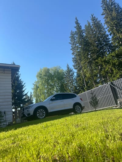 This media file presents a bright, outdoor scene, likely in Areyskoye, Russia, under a clear, sunny sky. The image is composed of a green lawn, a white car, part of a wooden house, a slatted fence, and a dense stand of trees.

In the foreground, a lush, vibrant green lawn spans the lower left, illuminated by strong sunlight that casts distinct shadows. A white car, possibly an SUV or crossover, is parked on the right side, partially visible behind the grass. The car is equipped with alloy wheels, one of which clearly displays a BMW logo on its hubcap. The license plate is partially obscured and its text is illegible.

Behind the car, in the mid-ground, a dark grey or black slatted fence extends horizontally across the frame. To the upper left, a section of a white-painted wooden building, likely a house with horizontal log-style siding, is visible, topped by a dark roof overhang.

The background is filled with numerous tall trees, primarily dark green coniferous varieties like pines or spruces, interspersed with lighter green deciduous trees. Thin black wires, possibly utility lines, are visible stretching across the canopy of the trees. The sky above is a brilliant, cloudless blue, indicating clear weather conditions. The strong illumination and shadows suggest the time of day is late morning or early afternoon. No individuals or specific activities are depicted, giving the scene a peaceful and serene ambiance in what appears to be a rural or suburban residential setting.