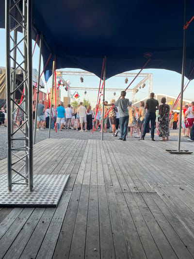 The image shows a crowd of people gathered under a large blue tent, possibly at an outdoor event or festival. The foreground features a wooden deck with metal scaffolding. In the background, there's a stage with lights and flags. The people are casually dressed, and some are standing or walking around. The overall scene suggests a lively atmosphere. The presence of flags suggests a celebration or international event. The weather appears to be clear and sunny.