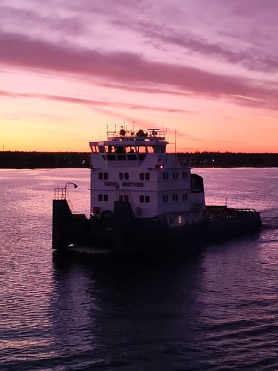 A river vessel is underway on a body of water at sunset. The sky is a mix of pinks, purples, and oranges. The water reflects the colors of the sky. The vessel has a white superstructure with windows. The name "ПАВЛИК ВИНОГРАДОВ" is visible on the side of the structure. The vessel is leaving a wake behind it. The opposite bank is a low, dark line of trees. Location: probably the city of Vichelovo, Russia.