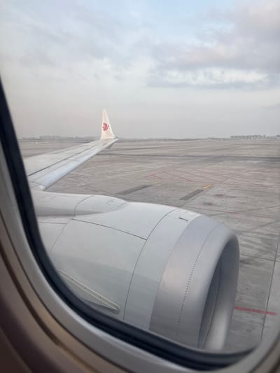 The image depicts an exterior view from inside an aircraft, looking through a circular passenger window. The primary subject is the starboard wing and engine of a China Eastern Airlines aircraft, visible in the foreground and midground. The engine cowling, primarily silver-grey, occupies the lower right quadrant, with its intake facing the viewer's right. The wing extends diagonally from the lower left, terminating in a visible wingtip featuring the red swallow-like logo of China Eastern Airlines. The aircraft is stationary on a concrete airport tarmac, which displays various faded red and yellow markings, including dashed lines and rectangular outlines. In the distant background, a low-rise city skyline is discernible against an overcast or hazy sky. The scene is located at Ürümqi Diwopu International Airport, Ürümqi, China. No people or active interactions are visible.