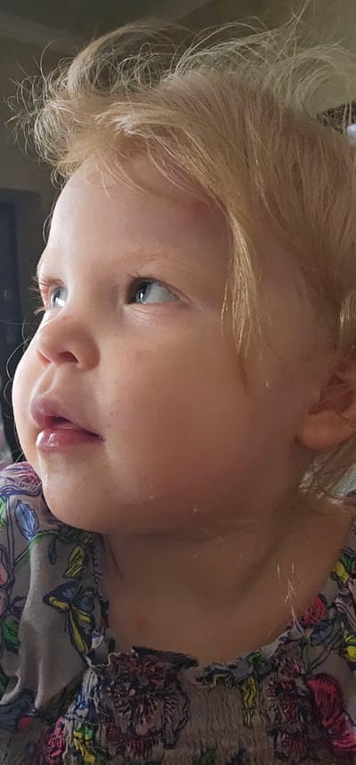The media file is a close-up, sideways photograph of a young child with blonde hair. The child's face is the primary subject, with their eyes looking upwards and to the left. Their lips are slightly parted, revealing a glossy appearance. The child is wearing a garment with a floral pattern. The background is out of focus and appears to be indoors, with some hints of natural light. There is no visible text in the image. The setting is not clearly identifiable beyond being indoors. The overall mood conveyed by the child's expression is neutral or contemplative. The image appears to be taken during the daytime.