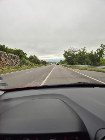 The image captures a view from inside a vehicle driving on a two-lane asphalt road. The road curves slightly to the right and stretches into the distance under a cloudy, overcast sky. On the left side of the road, a rocky hillside covered with lush green trees and vegetation rises sharply. On the right side, a metal guardrail runs parallel to the road, separating it from more trees and a gentle slope. A few vehicles are visible ahead on the road, appearing as dark shapes in the distance. The foreground shows the dashboard of the vehicle, with a portion of the windshield and a glimpse of the instrument cluster. The location is identified as being in the vicinity of Bogyovtsi, Bulgaria, suggesting a rural or mountainous setting.