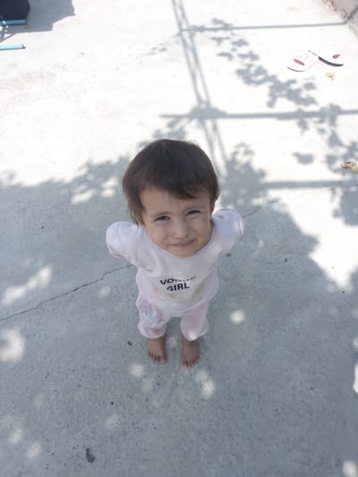 A young child, appearing to be a toddler, stands on a concrete surface outdoors. The child is wearing light pink pajamas with a graphic design on the front that includes the words "Vogue Girl". The child has dark hair and is looking up at the camera with a slight smile. The concrete surface shows signs of wear and tear, with visible cracks and some dark spots. In the upper right portion of the image, a pair of pink child-sized slides are visible. Shadows from what appears to be tree foliage are cast across the concrete. The environment suggests a residential outdoor area, possibly a courtyard or patio, in the city of Gagarin, Uzbekistan.