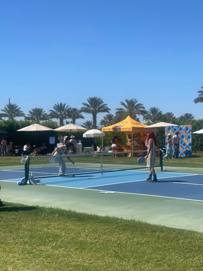 Here is a description of the image:

The image depicts an outdoor scene, likely a pickleball or tennis tournament in Thermal, California. A blue and green court is visible in the foreground, with a green grassy area on one side. People are visible playing and watching. Tents are set up along the side of the court, providing shade and vendor space. Palm trees line the perimeter of the area against a clear blue sky, indicating a sunny, warm climate.