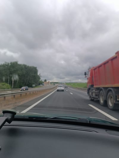 The image depicts a view from the interior of a vehicle on a highway, looking forward. The sky is overcast with dense, grey clouds. The highway has multiple lanes, with white lines marking the boundaries. In the foreground, the dashboard of the vehicle is visible, along with a suction cup mount on the windshield.
Several vehicles are on the highway. To the right, a large red dump truck is driving in the same direction as the camera vehicle. In the lanes to the left, a white passenger car is visible, and further ahead, another car is slightly blurred. The highway is bordered by a metal guardrail on the left, and beyond that, lush green trees form a dense backdrop. In the distance, there are hints of buildings or structures. The overall impression is of a journey on a highway under cloudy weather conditions, likely in a rural or semi-rural area. The location is identified as the Versyališčawski sel'ski Savec in Belarus.