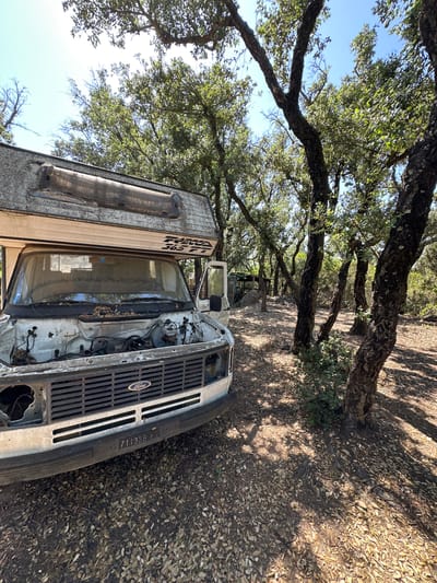 An old, white Ford camper van, likely a Ford E-Series based RV, is parked in a wooded area. The van is heavily weathered and appears to be abandoned or in disrepair. The hood is open, revealing the engine compartment, and the front grille is damaged. Leaves and debris are scattered on the ground and on the van's surfaces. The camper body, visible above the cab, has "FRECCA 365 FT" lettering. The vehicle's license plate reads "711359 HG". The surrounding environment consists of trees with rough bark and green foliage, casting dappled shadows on the ground which is covered with fallen leaves. The sky is clear and blue. The scene appears to be in a rural or semi-rural setting, potentially on the outskirts of Santa Cristina d'Aro, Spain, given the oak-like trees and the style of the license plate. There are no people visible in the image.