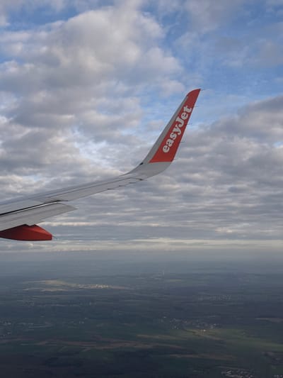 The image shows a view from an EasyJet airplane window, with the wing visible in the foreground. The wing has the "easyJet" logo printed on it in white on an orange background. Beyond the wing, there is a sky filled with clouds, with some patches of blue visible. In the background, a landscape is visible, appearing to be a mix of green fields and urban areas, possibly near the city of Berg in Luxembourg. The land is partially obscured by a layer of haze or fog. The photograph is oriented with the wing on the left side of the image and the landscape on the right.