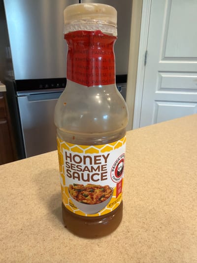 The image shows a bottle of Panda Express Honey Sesame Sauce standing on a speckled countertop. The bottle is mostly clear, with a light brown liquid inside. The label is predominantly white and yellow, featuring the words "HONEY SESAME SAUCE" in large, bold, dark letters. A smaller circular logo with a panda and the words "PANDA EXPRESS CHINESE KITCHEN" is also visible. A picture of a serving suggestion, showing chicken and vegetables in the sauce, is present on the lower part of the label. A red band with repeating text, likely the brand name, wraps around the neck of the bottle. In the background, a stainless steel refrigerator and a white door are partially visible. The scene appears to be indoors, possibly in a kitchen. The provided context states the location is Carpentersville, United States of America.