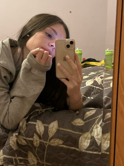 A young woman takes a selfie in a mirror, holding her phone close to her face. She wears a gray zippered hoodie. Her hair is dark, and her expression is neutral. The background includes a bed covered with a gray and white floral-patterned quilt. A small fly is visible in the upper left of the mirror.