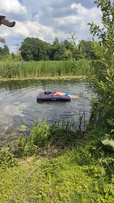A young male wearing blue swim trunks is lying on a blue inflatable mattress in a body of water. He is face down, with his head turned to the left, and his arms extended. The mattress has the brand name "Bestway" visible on it. The water is calm, with ripples emanating from the mattress. Lily pads are visible on the surface of the water, especially in the foreground and to the left of the mattress. The background consists of tall green reeds lining the bank of the water, followed by a dense forest of green trees. Above the trees, a cloudy sky is visible. To the left side of the image, a wooden structure with a curved edge is partially visible. The scene appears to be a natural outdoor setting, possibly a river or lake, in Ramon, Russia.