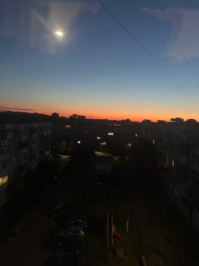 The image depicts a twilight view of a residential area in Baltiysk, Russia. The sky transitions from a deep blue at the top to a vibrant orange and pink horizon, indicating sunset or sunrise. In the sky, a bright light, possibly a street lamp or reflected moonlight, is visible, along with what appears to be a small, distant star or planet. Thin power lines stretch across the upper portion of the frame.

Below the sky, multi-story apartment buildings with illuminated windows dominate the scene. The buildings are characterized by their block-like architecture and balconies. The lower ground level shows a courtyard area with a playground consisting of a slide and climbing structure. Several parked cars are visible in the dark courtyard, with some headlights on. Trees and shrubbery cast dark shadows across the scene, adding to the low-light ambiance. The overall environment suggests a quiet evening in a typical urban residential setting.