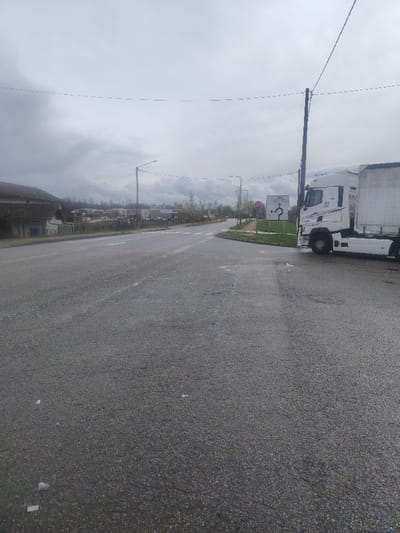 The image displays a wet asphalt road under an overcast sky at a junction in Servas, France. A white Mercedes-Benz Actros semi-truck and its trailer are stationary on the right shoulder, adjacent to a grassy median. This median features a red circular STOP sign and a white square directional sign indicating a roundabout for 'Servas,' 'St Paul de Varax,' and 'Meillonnas.' A wooden utility pole with overhead wires stands behind the truck. On the far left, a segment of a building with a terracotta-tiled roof is visible. Streetlights and trees with light green foliage line the road, which recedes towards distant structures under the cloudy expanse. No individuals are present, and no actions are occurring.