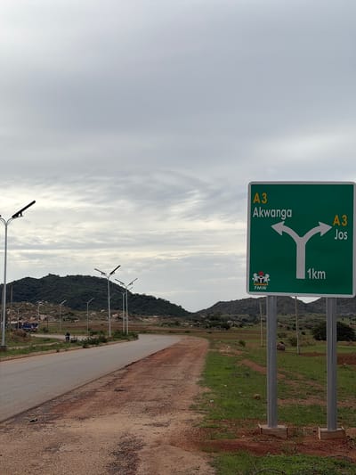 The image depicts a road sign in Foram, Nigeria, indicating directions to Akwanga and Jos. The sign, which is green with white and yellow lettering, shows "A3 Akwanga" with an arrow pointing left, and "A3 Jos" with an arrow pointing right. Below the arrows, "1km" is displayed. In the bottom left corner of the sign, a coat of arms with "FMW" written underneath is visible. The sign is mounted on two metal poles. The scene shows a paved road curving to the left, with a dirt shoulder on the right. In the background, there are green hills covered with vegetation, and several lampposts line the side of the road. The sky is overcast with a cloudy, gray appearance. There are no people prominently featured in the image, but a person on a bicycle can be seen in the middle distance on the left side of the road.