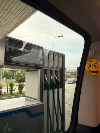 This is a medium-shot, rotated image taken from inside a vehicle, looking out through the windshield at a gas station. The scene is set in Podlugovi, Bosnia and Herzegovina.
The foreground is dominated by the interior of the car, with the grey headliner and the black frame of the windshield visible. A yellow car air freshener with a smiley face and the word "areon" is hanging from the roof. Reflected in the windshield is a gas pump with multiple nozzles. The pump displays labels for different fuel types, including "DIZEL" and "PREMIUM 95," as well as payment options like "VISA." There's also text indicating "KM/L" and "IZDANO LITARA," suggesting fuel dispensing measurements.
Through the windshield, the gas station itself is visible, along with vehicles parked at the pumps. Beyond the gas station, there are buildings and trees, indicating an urban or semi-urban setting. A street lamp is also visible in the distance. The sky is bright and appears to be daytime, with a slightly overcast or hazy quality. The overall impression is a mundane stop at a gas station.