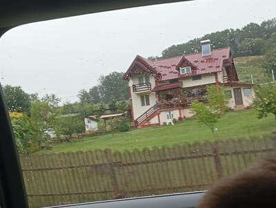 A scenic view from inside a vehicle captures a multi-story house with a red tiled roof and white and tan siding, situated on a grassy hillside. The house features multiple windows, balconies, and a prominent staircase on its exterior. A wooden fence runs horizontally across the foreground. Trees and foliage surround the property, with a cloudy, overcast sky above, suggesting a rainy day as evidenced by the water droplets on the vehicle's window. The location is identified as Curtea de Argeș, Romania.