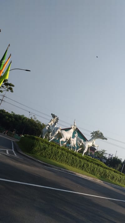 In Trosobo, Indonesia, a statue of white horses is displayed on a green, manicured median. The statue features several horses in various dynamic poses, as if in mid-gallop. The horses are made of a white, possibly marble or plaster, material. The median is bordered by a road with white lane markings. In the background, a building with a green roof and light-colored walls is visible, along with trees and power lines. Several colorful flags are also present in the upper left portion of the image, and a bird is seen flying in the clear blue sky. The scene appears to be outdoors, under a bright sky, suggesting it is daytime.
