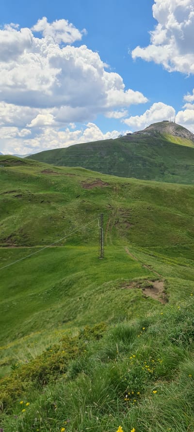 The image depicts a vibrant, green mountainous landscape under a bright blue sky dotted with fluffy white clouds. The terrain is characterized by rolling hills covered in lush grass, interspersed with patches of darker earth and scattered yellow wildflowers in the foreground. A prominent metallic pole, part of what appears to be an overhead cable system, stands in the middle ground, with a thin cable stretching diagonally across the frame.
In the background, the slopes of a larger mountain rise, featuring a series of terraced green areas, possibly for agriculture or erosion control, demarcated by horizontal lines of vegetation and wooden posts. The summit of this mountain is capped with structures, including buildings and at least two antenna towers, suggesting a ski resort or a scientific installation. The overall scene conveys a sense of natural beauty and open space, typical of alpine regions. The location is identified as Sëlva - Wolkenstein - Selva di Val Gardena, Italy.