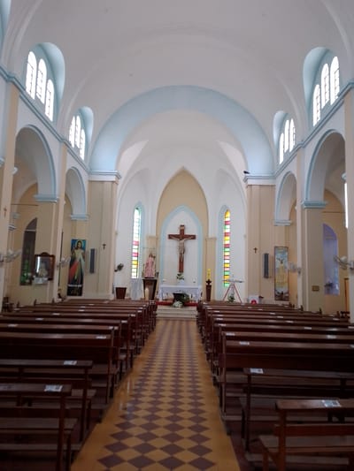 The image shows the interior of a church. Rows of wooden pews are arranged on either side of a checkered aisle. At the far end is an altar with a crucifix. Stained glass windows are visible. The building has an arched ceiling and multiple arched windows. A poster is visible on the left side of the church. The church is located in the city of Villa Hayes, Paraguay.