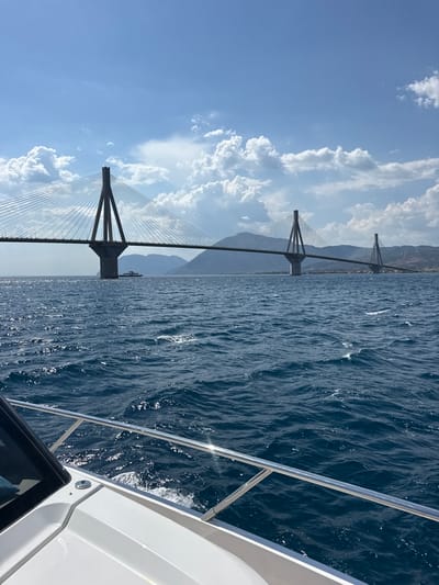 This is a photograph taken from a boat, looking towards a large cable-stayed bridge spanning a body of water. The foreground of the image shows the white hull and a chrome railing of the boat, with the dark blue water extending outwards. The middle ground is dominated by the expansive bridge, supported by tall concrete pylons, with numerous cables stretching to the roadway. A small ferry or boat can be seen underneath the bridge. In the background, mountains rise, and a hazy coastline is visible, with what appear to be wind turbines on a distant hill. The sky is a bright blue, dotted with white, fluffy clouds. The scene is likely the Rio-Antirrio Bridge, which connects the Peloponnese peninsula to mainland Greece, crossing the Gulf of Corinth near the city of Rio in the Municipal Unit of Rio, Greece. The overall impression is one of a bright, sunny day on the water, with a focus on the impressive engineering of the bridge.