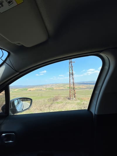 The image captures a rural landscape viewed from the passenger-side interior of a vehicle on a clear, sunny day. The upper and lower portions of the frame are occupied by the vehicle's interior: a grey sun visor and headliner above, and a dark grey door panel with a chrome-finished handle below. A black cable runs vertically along the left A-pillar. The exterior side mirror on the left reflects a section of an asphalt road and a light-colored vehicle.

Outside the window, a expansive environment unfolds under a blue sky dotted with scattered white clouds. In the midground, a tall, rusty, metal lattice power transmission tower stands on a grassy incline, with power lines extending from its structure. The surrounding terrain consists of green and brown agricultural fields. In the far background, a cluster of buildings comprising the settlement of Razboyna, Bulgaria, is visible, extending across the landscape to meet distant, undulating hills. No individuals or specific actions are discernible within the frame.