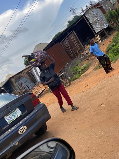 In Rahwol Chom, Nigeria, a woman carries a large bundle on her head, balanced by her right hand. She wears a black jacket and red plaid pants, with grey sandals. A dark blue car is parked in the foreground, its license plate reading "BLL 508TG". To the right, a young man in a blue shirt and patterned pants stands with his hands in his pockets, looking up. In the background, corrugated metal structures and wooden planks suggest a market or workshop area. Power lines stretch across the sky, hinting at the infrastructure of the environment. The ground is a dusty, reddish-brown earth.