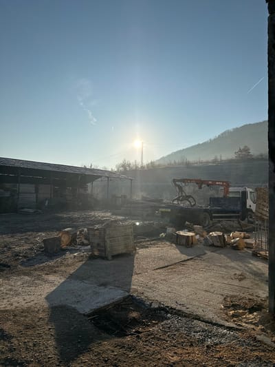 The scene depicts an outdoor industrial yard in Dobrodan, Bulgaria, under a clear, light blue sky with a low-angle sun, suggesting either early morning or late afternoon. The environment appears cold, indicated by visible mist or frost on the distant treed hillside.
In the foreground, the ground is a mix of packed dirt and gravel, with an exposed section of rebar mesh within a muddy depression. Long shadows are cast from left to right across this area.
The midground features an operational area. To the left, a large, open-sided shed with a corrugated metal roof houses stacks of timber or planks. In front of the shed, a large, cut log section rests on the ground, and a wooden pallet stacked with materials is centrally positioned. To the right of center, a white flatbed truck equipped with an orange articulated crane, marked "ATLAS 3006" and featuring a timber grab attachment, is parked. Several raw log sections are scattered on the ground near the truck.
The background shows a rolling hillside covered with trees, partially obscured by a light atmospheric haze or frost. A utility pole with power lines is visible against the hillside, near the bright sunburst. No individuals are present in the frame. The overall impression is that of a timber yard or similar industrial facility engaged in the processing or handling of wood.