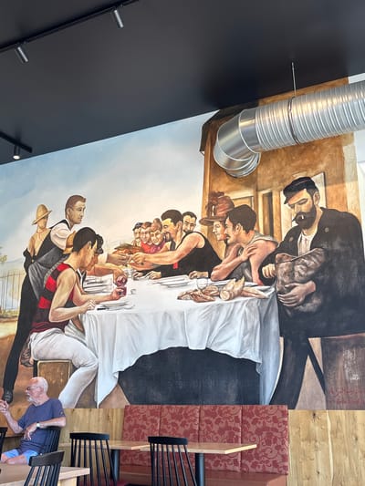 This is a photograph of a mural painted on a wall, likely in a restaurant or dining establishment in Elsenfeld, Germany. The mural depicts a scene of people gathered around a long table, reminiscent of a feast or communal meal. The style of the painting is realistic with a warm, earthy color palette.
The mural features a diverse group of individuals, painted in various states of engagement with each other and the food laid out on the table. Some figures are leaning in, talking or sharing food, while others appear more pensive. The table is laden with bread and other dishes, covered partly by a white tablecloth. The people are dressed in what appears to be historical or period clothing, suggesting a narrative or thematic representation.
Notable details include the artistic signature "A.Sultan 2023" and what looks like "by Emilo Triuggi" also visible on the wall. The background of the mural transitions from a darker, more somber tone at the top to a lighter, sky-like blue towards the bottom, possibly indicating an outdoor setting or a progression in the narrative. The photograph also captures architectural elements of the space, including exposed metal ductwork, spotlights, and upholstered seating with a floral pattern, all hinting at a contemporary interior design contrasting with the historical depiction in the mural. The overall mood is one of communal gathering and shared experience.