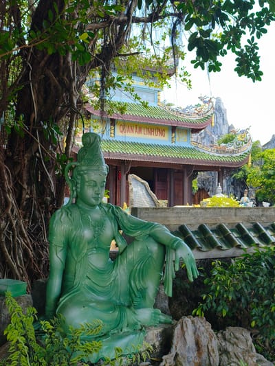 A bright, eye-level, medium shot captures a vibrant green jade statue of a seated deity, partially obscured by the thick, gnarled roots of a large tree on the left. The statue, made of smooth, polished jade, depicts a figure with serene features, elaborate hair ornaments, and flowing robes. Its right hand is extended, fingers splayed outwards in a gesture of offering or blessing. The background reveals a traditional Vietnamese temple with a distinctive green tiled roof, adorned with ornate carvings and a golden inscription that reads "QUAN ÂM LINH ỨNG". Towering limestone karst formations rise majestically behind the temple, their rugged textures contrasting with the intricate architecture. Lush green foliage surrounds the scene, with trees and plants creating a sense of natural beauty and tranquility. The overall atmosphere is one of peace and spiritual reverence.
