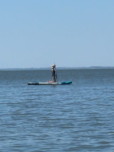 A person wearing a wide-brimmed hat and a black swimsuit kneels on a paddleboard in the water. The paddleboard is a light gray color with a blue section at the front. The person is holding a paddle upright. The water is a deep blue with small ripples. In the distance, a faint landmass with trees is visible against a clear blue sky. The scene appears to be a calm day on a body of water.