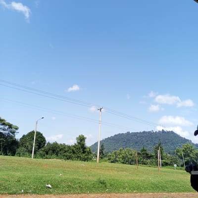 The image depicts a landscape in Ihururu, Kenya, under a blue sky with scattered clouds. In the midground, a grassy field slopes upwards towards a line of trees and bushes. Power lines stretch across the scene supported by utility poles. A mountain covered with dense forest rises in the background. A street lamp and some wooden posts are scattered in the area. Part of a person is to the right.